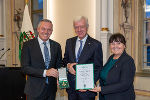 Bildbeschreibung: Drei Personen in einem Raum. Sie halten eine Urkunde und ein Ehrenzeichen in den Händen.