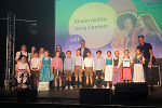 Gruppe von Kindern und Jugendlichen auf einer Bühne, die Preise entgegennehmen. Im Hintergrund eine Wand mit dem Schriftzug 'Kindercontest seit 2019'.