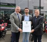 Bildbeschreibung: Drei Personen bei einer feierlichen Übergabe von einer Urkunde und Blumen vor einem Motorradgeschäft.