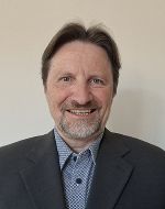 Porträt einer Person mit grau meliertem Haar und Bart. Die Person trägt einen dunklen Anzug und lächelt in die Kamera.