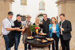 Gruppe von Menschen steht um einen Grill und hält Spieße mit Fleisch und Gemüse. Im Hintergrund ein Gebäude.