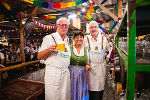 Bildbeschreibung: Drei lächelnde Personen in Tracht mit Bierkrügen auf einem Volksfest. Im Hintergrund ein festlich geschmücktes Zelt.