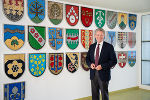 Bildbeschreibung: Ein Mann im Anzug steht vor einer Wand, an der viele Wappen hängen.