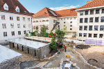 Bildbeschreibung: Bauarbeiten in einem Innenhof mit Personen, einem Kran und Bepflanzungen zwischen weißen Gebäuden.